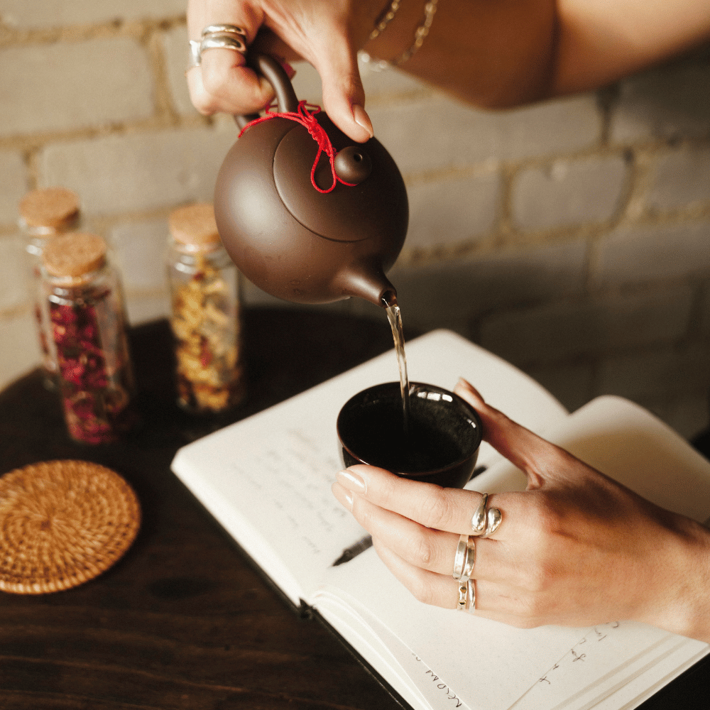 Inner Code tea ritual. A person pouring hot water from a dark brown ceramic teapot with a red cord tie into a small black cup, set on a dark table with an open notebook, rings, and glass jars of dried flowers in the background.